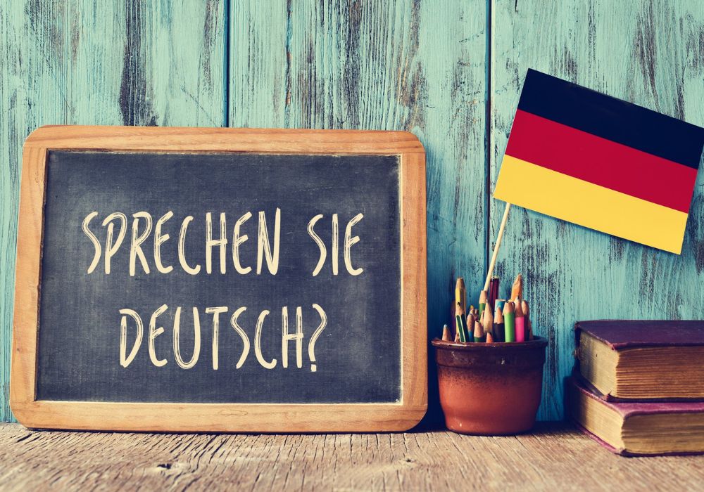 Sur la photo il y a une ardoise sur laquelle est écrit "Sprechen Sie Deutsch?" (Parlez-vous allemand?). A côté de l'ardoise sont un drapeau allemand, des stylos et deux livres.